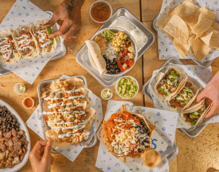 Overhead view of hands reaching for trays of tacos, burritos, chips with salsa, and side dishes on a wooden table.